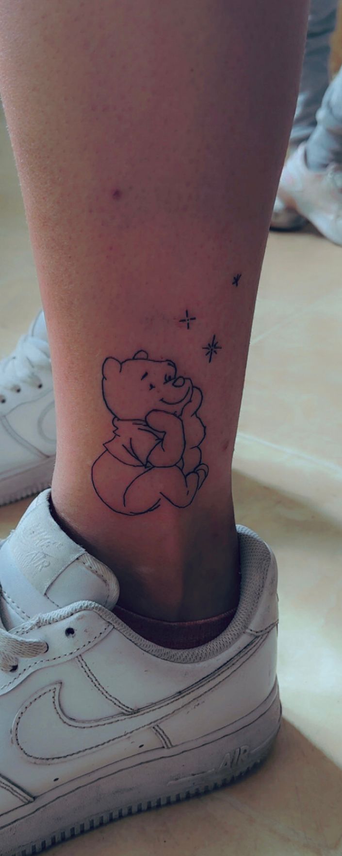 Tatouage fineline Winnie l'Ourson assis avec etoiles cheville Hakuna Tattoo